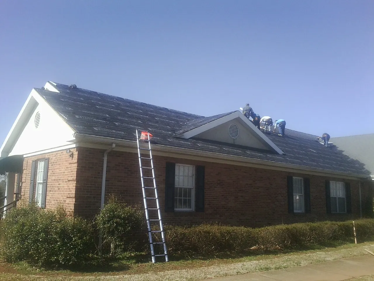 Crew tearing shingles off a brick ranch roof with a ladder set — Emergency Roof Repair work by our crew in Deansboro