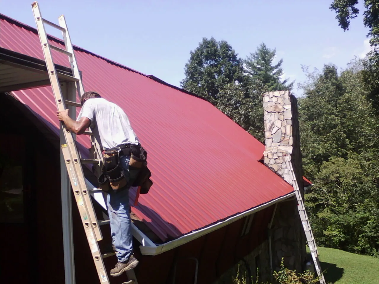 Roofer on a ladder beside a red metal roof and stone chimney — Soffit & Fascia Repair by Roofing Brothers in Deansboro